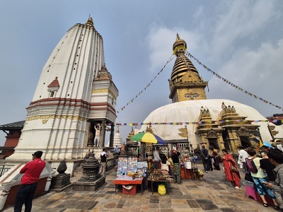 �irokouhl� z�ber na hlavn� budhistick� st�pu Swayambhunath s pozl�ten�m ihlanom a nama�ovan�mi Buddhov�mi o�ami vpravo. V�avo dominuje biela ve�a Pratappur v �t�le Shikhara. Medzi nimi prebieha �ul� ruch � n�v�tevn�ci obch�dzaj� st�pu a v popred� stoj� st�nok so spomienkov�mi predmetmi.