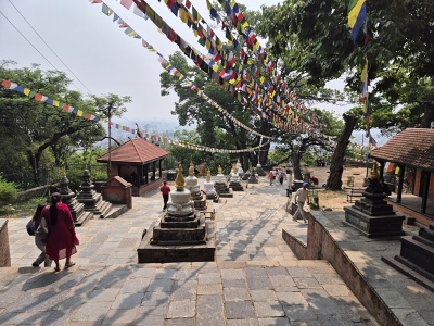 Poh�ad smerom nadol po �irokom kamennom schodisku s 365 schodmi v komplexe Swayambhunath. Nad schodiskom s� natiahnut� farebn� budhistick� modlitebn� vlajo�ky a po bokoch st�py stoja mal� biele a kamenn� chorteny. V pozad� presvit� panar�ma mesta K�thm�ndu a stromy obklopuj�ce kopec.