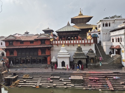 Poh�ad z opa�n�ho brehu rieky Bagmati na hlavn� hinduistick� chr�mov� komplex Pashupatinath v K�thm�ndu so zlatou strechou pagody, kamenn�mi sv�ty�ami a obradn�mi gh�tmi so schodiskami ved�cimi k vode.