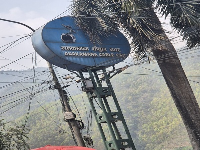 Ve�k� modr� okr�hla tabu�a s n�pisom Manakamana Cable Car a obr�zkom kab�nky, umiestnen� na sto�iari pred zelen�mi h�rskymi svahmi v Nep�le