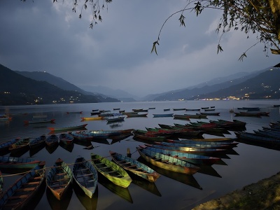 Farebn� dreven� veslice kotviace na pokojnej hladine jazera Phewa po�as s�mraku s rozsvieten�mi svetlami mestskej �asti Pokhara a siluetami h�r v pozad�.