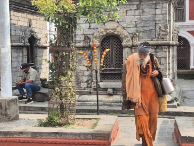 Star�� hinduistick� sv�t� mu� s�dhu s dlhou bielou bradou, v tradi�nom oran�ovom odeve kurta a dh�t� a s n�hrdeln�kmi rudr�k�a, kr��aj�ci po kamenn�ch n�dvoriach historick�ho hinduistick�ho chr�mov�ho komplexu v Nep�le