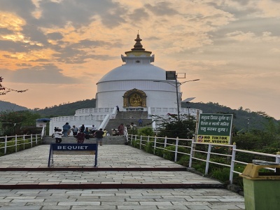 Biela pagoda World Peace Pagoda so zlatou sochou Budhu v podve�ernom osvetlen�, pr�stupov�m kamenn�m chodn�kom a dramatickou oblohou v Pokhare.