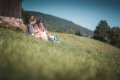 Dvojica sed� v tr�ve pri drevenej stavbe, ved�a nich le�� batoh